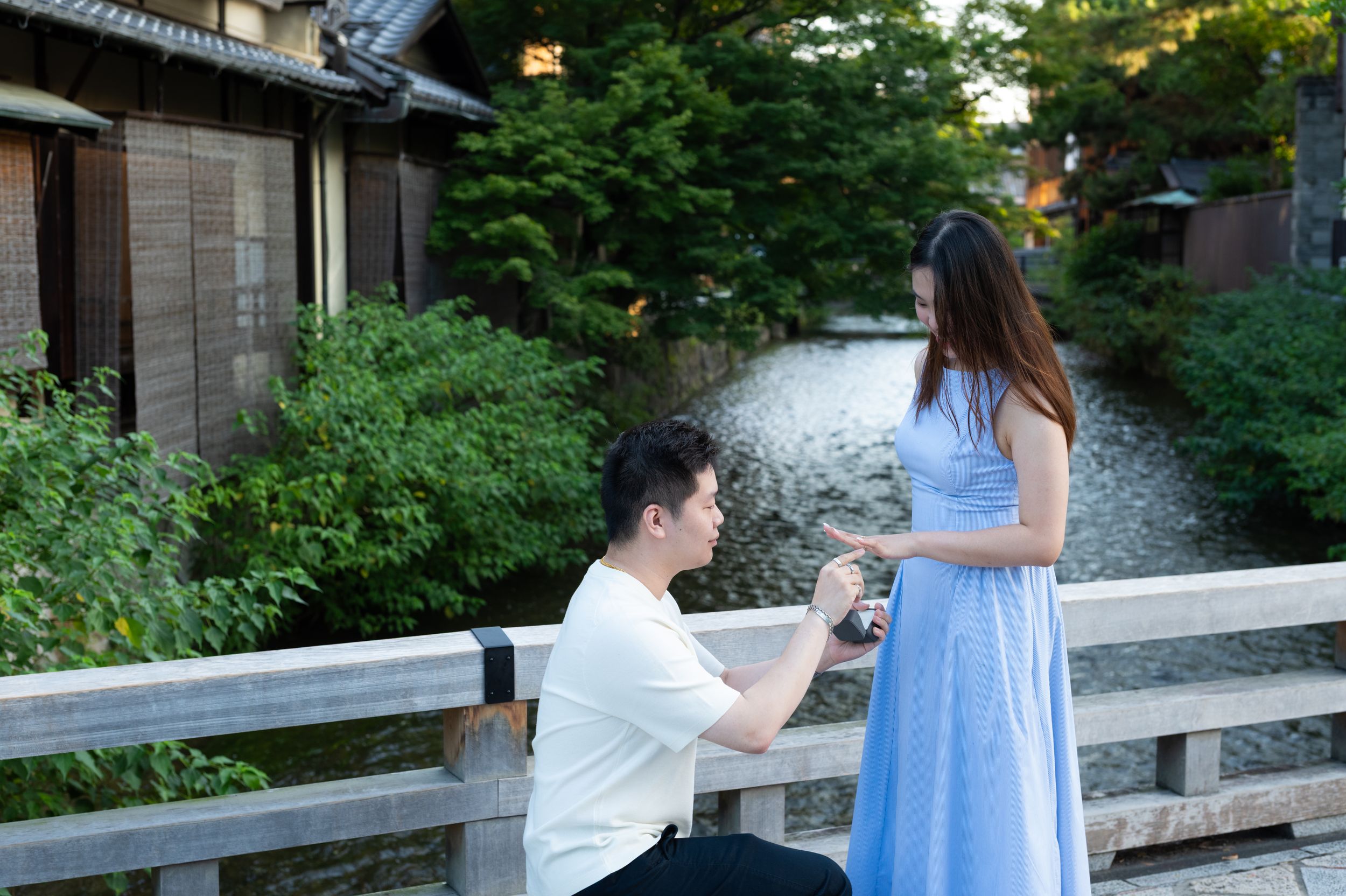 Man kneels on a wooden bridge proposing to a woman in a long blue dress, with a calm stream and lush green trees in the background.