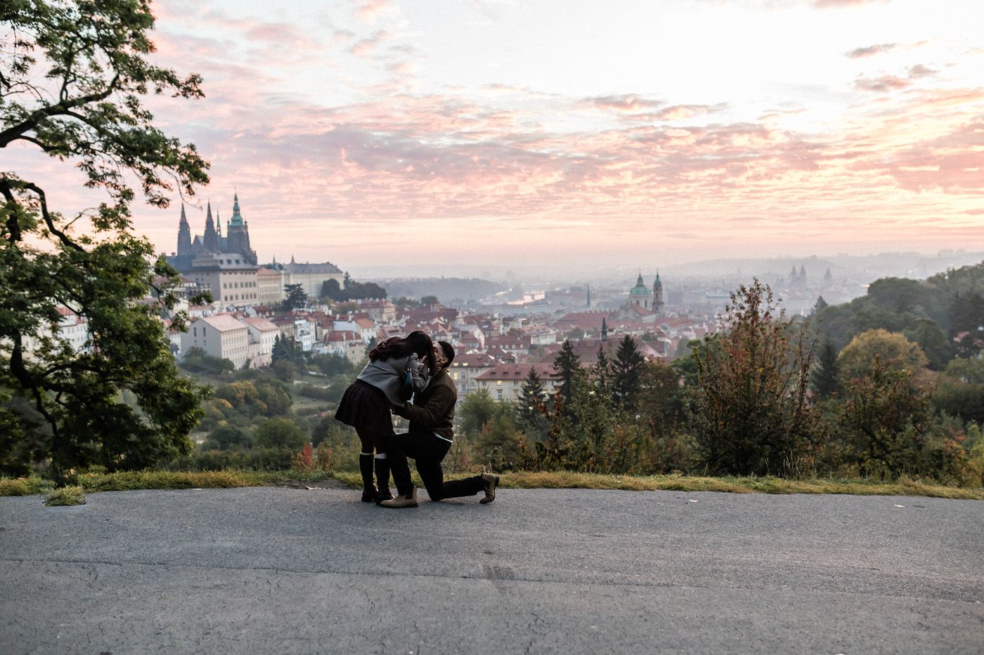 Couple shares a marriage proposal moment on a hilltop path at sunset, overlooking a historic city skyline with a cathedral and pink clouds.