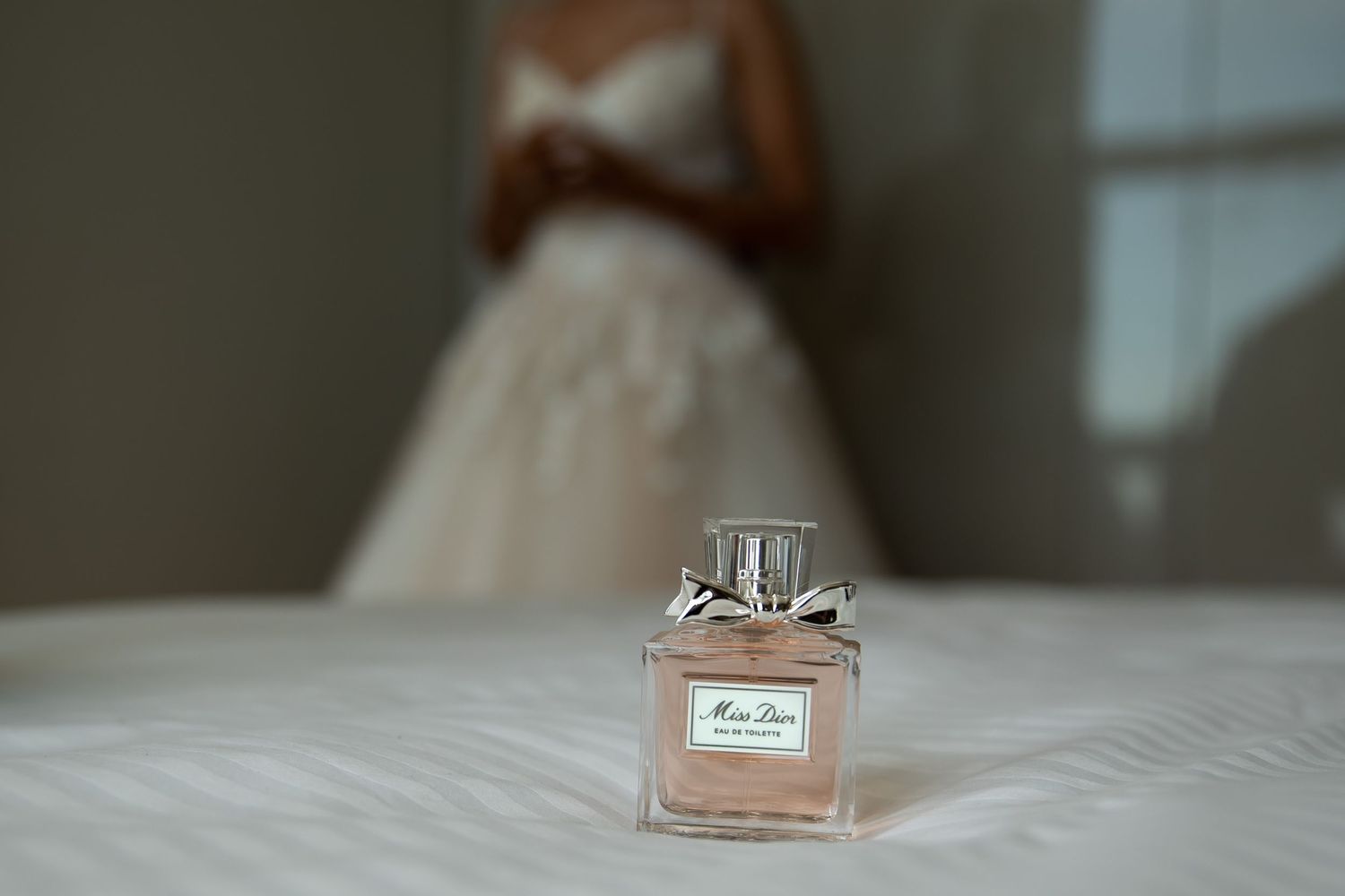 Miss Dior perfume bottle on white surface with blurred bride in wedding dress in background.