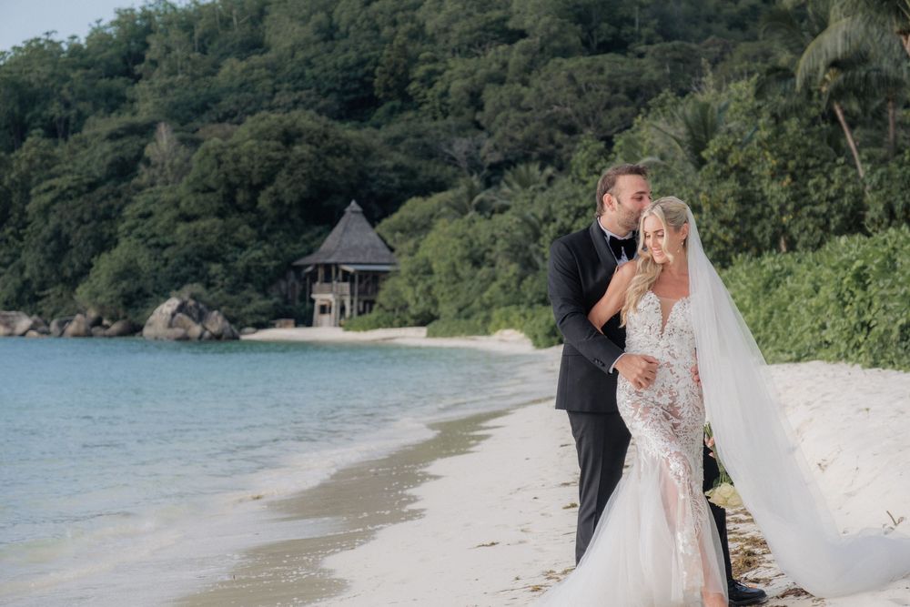Bride and groom share a romantic moment on a tropical beach with lush greenery and a gazebo in the background.