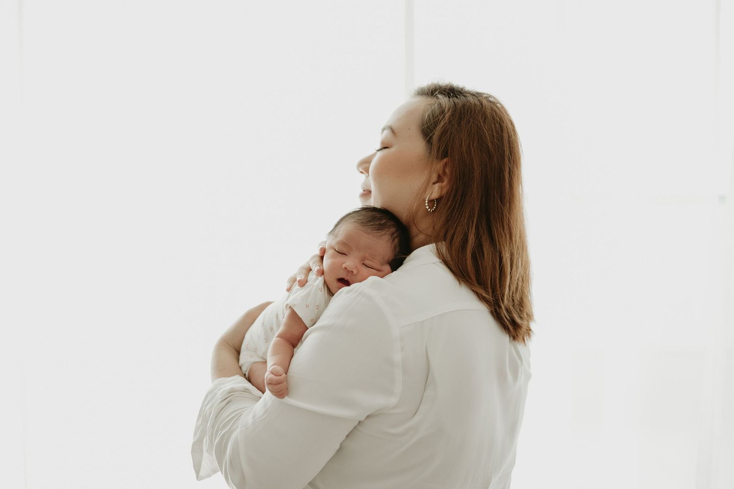 A smiling mother in a white shirt tenderly holds her sleeping newborn baby against her chest in a bright, airy room.