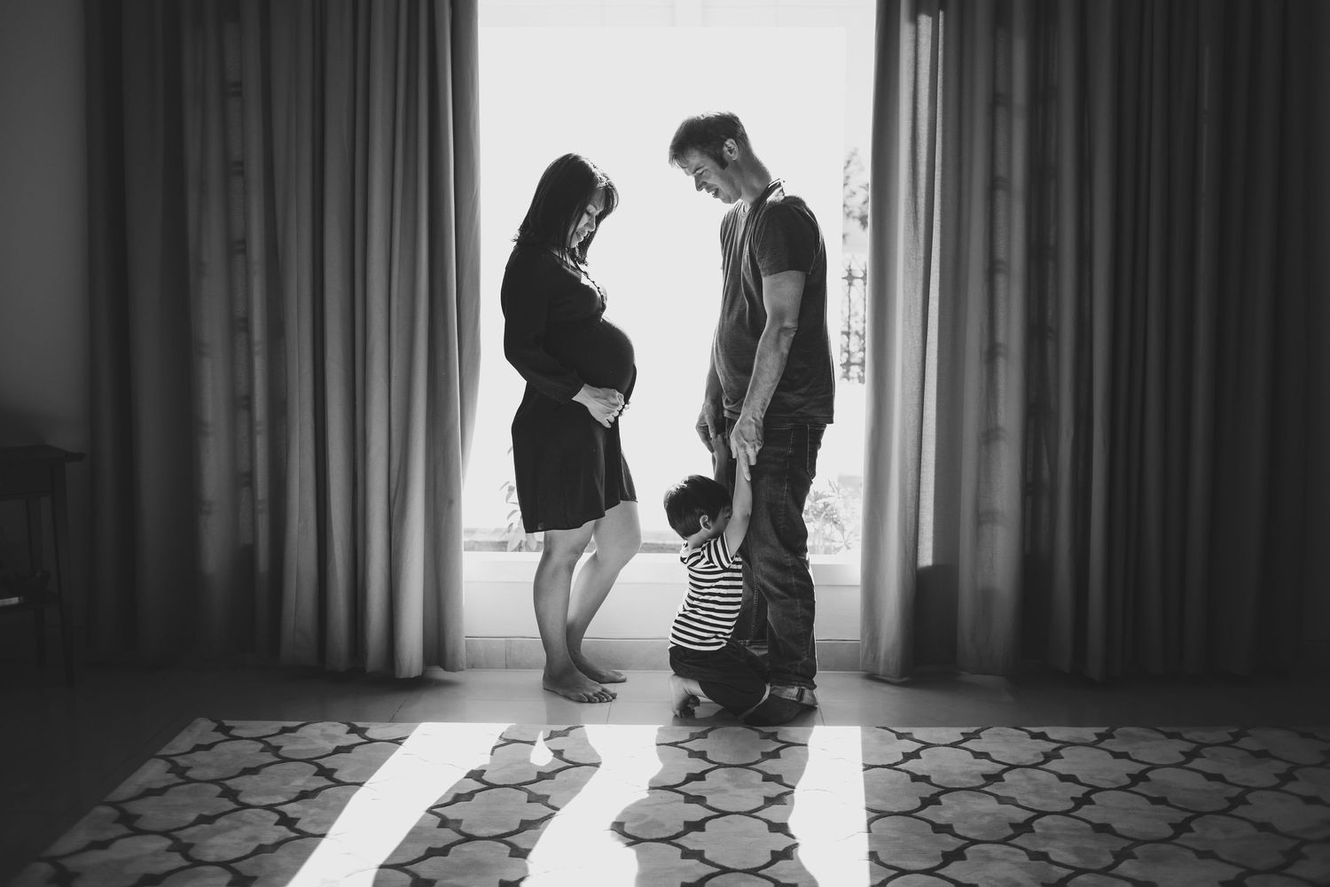 Black and white maternity photo of pregnant woman, man, and toddler standing by a sunlit window indoors.