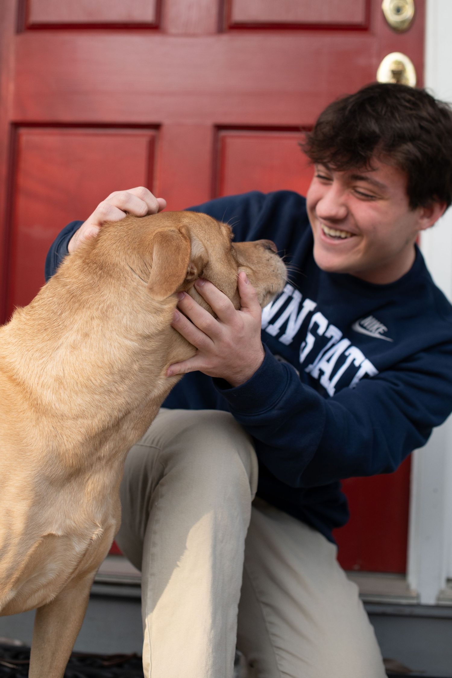 Young man in Penn State sweatshirt smiling while petting a golden dog near a red front door.