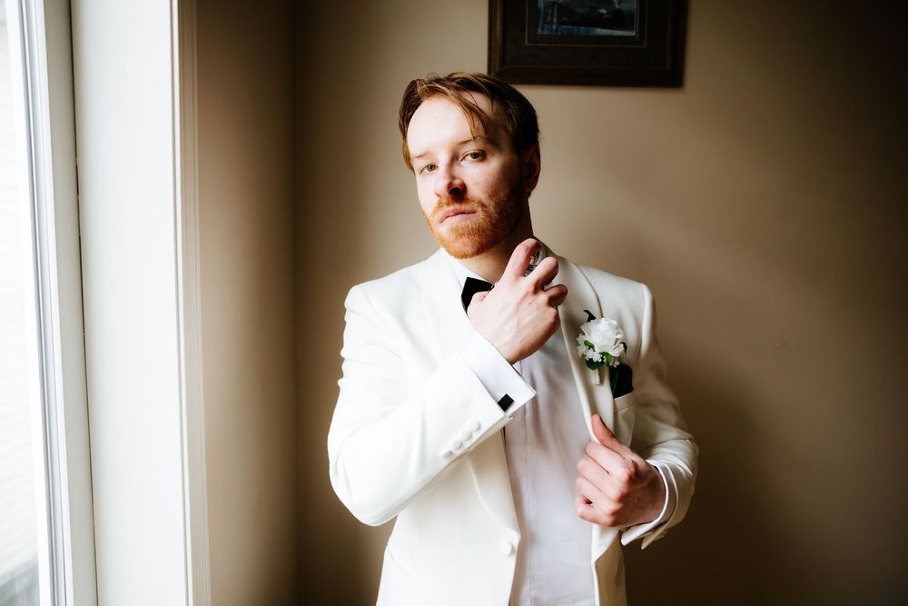 Groom in white tuxedo adjusting bow tie with boutonniere, standing by window in elegant wedding attire.