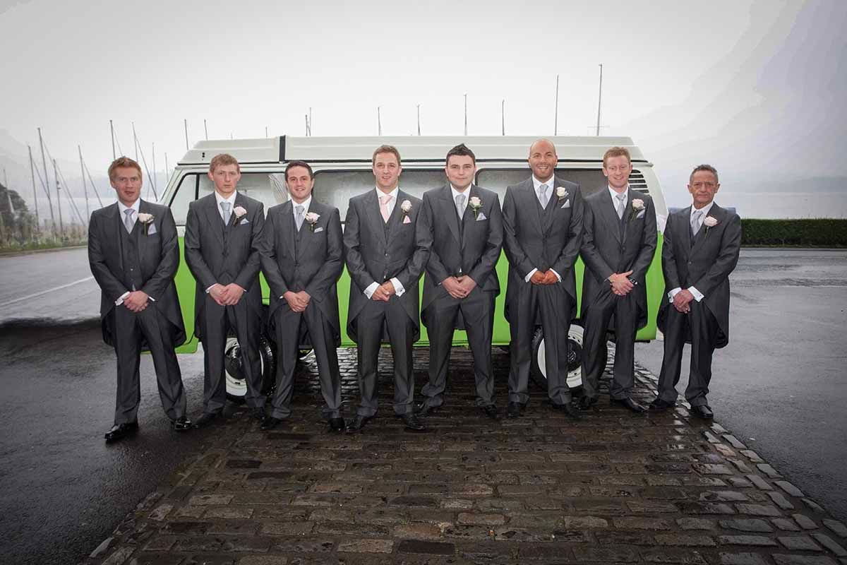 Seven groomsmen in grey suits pose in front of a green and white van on a cobblestone surface.