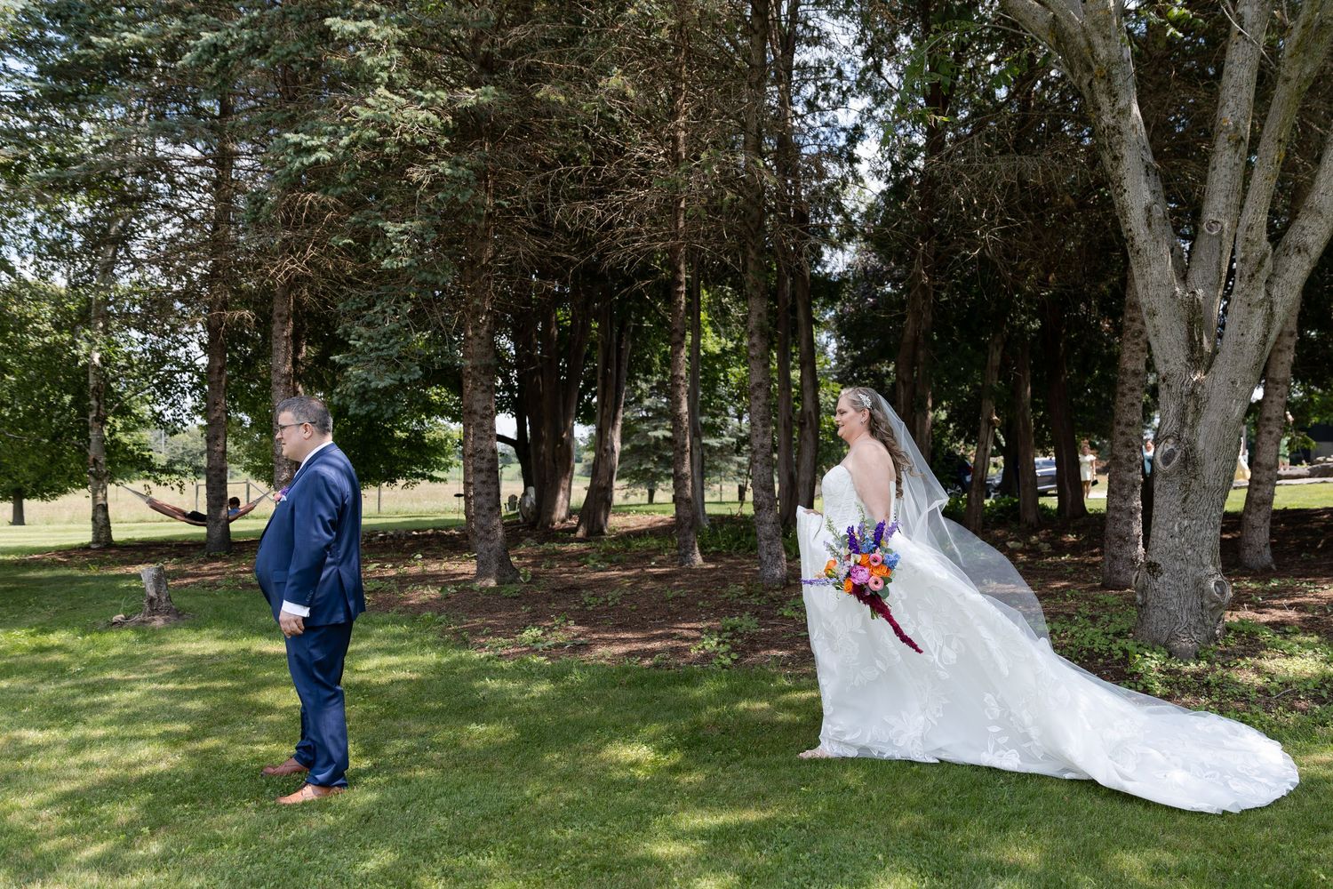 Bride in white gown with colorful bouquet faces groom in navy suit during outdoor first look near tall trees in Guelph, Ontario.