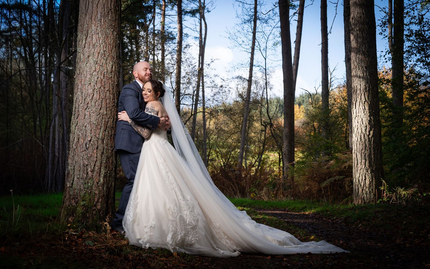 Bride in white gown and groom in suit embrace by tall trees in an autumn forest wedding portrait.