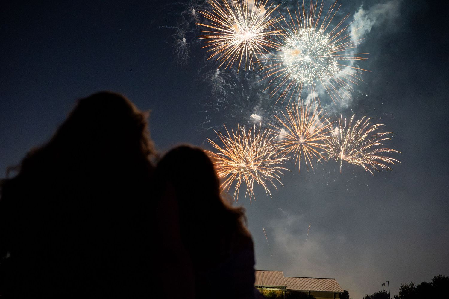 Two silhouetted figures watch colorful fireworks burst in a dark night sky.