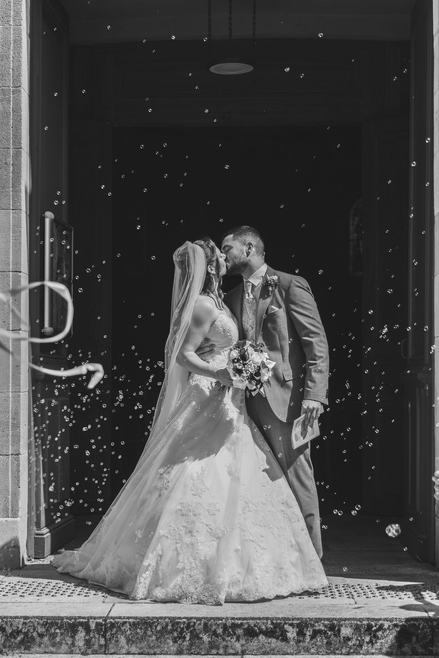 Black and white photo of a bride and groom holding hands, walking on grass in front of an elegant mansion.