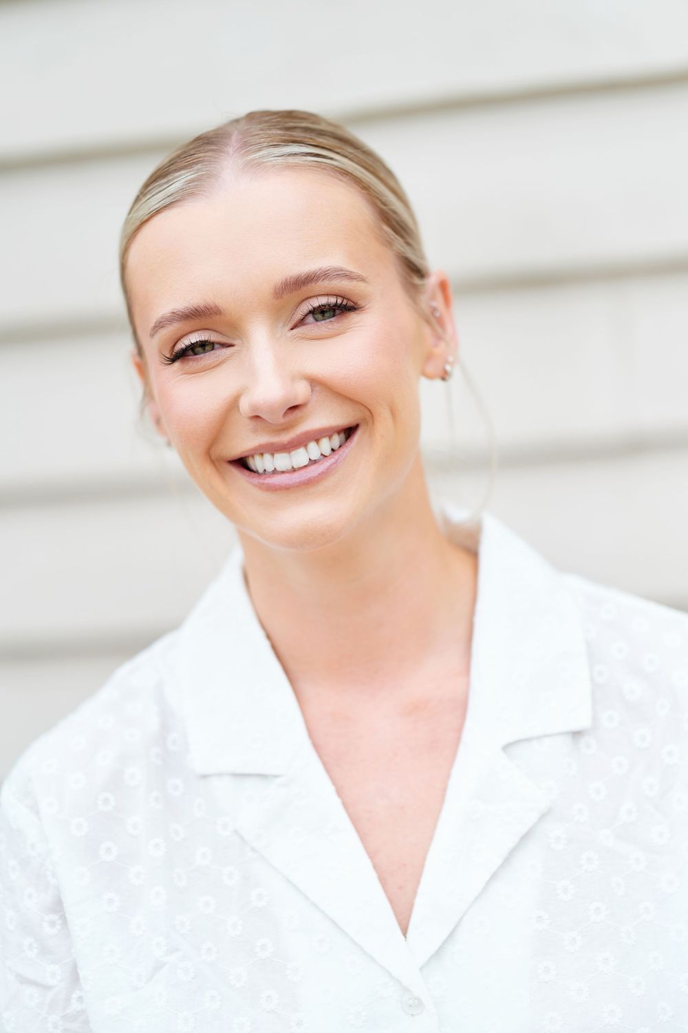 Smiling blonde woman with pulled-back hair wearing a white collared shirt, photographed outdoors against a light background.