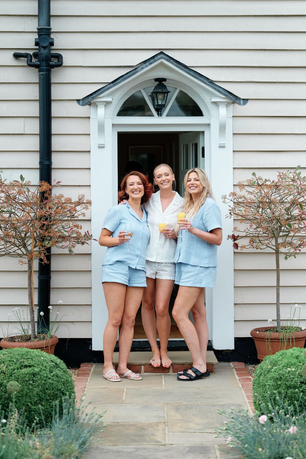 Three women in matching blue pajamas smile holding drinks outside a charming white house entrance.