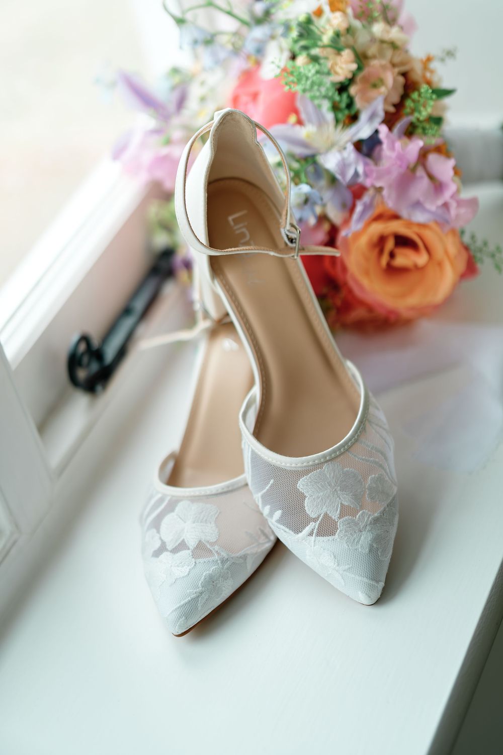 Elegant white lace bridal heels with ankle strap on windowsill beside a colorful wedding bouquet.
