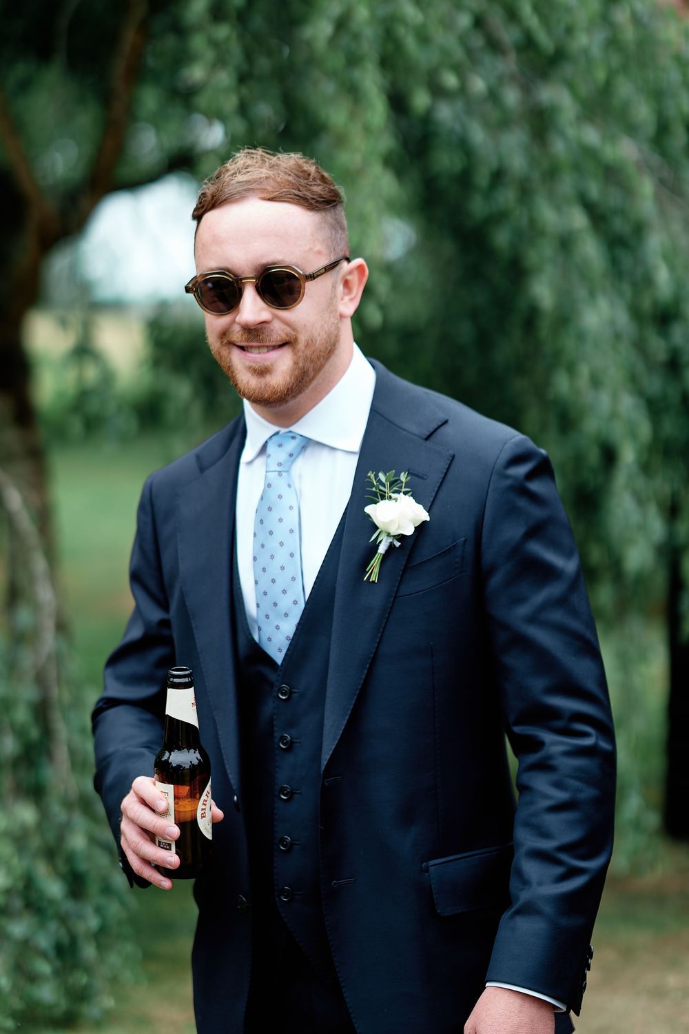 Smiling groom in navy three-piece suit with boutonniere, light blue tie, sunglasses, holding beer bottle outdoors.