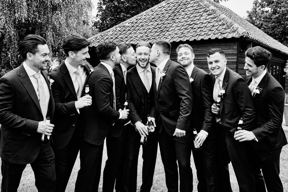 Black and white photo of a groom and groomsmen laughing together in suits outside a rustic barn venue.