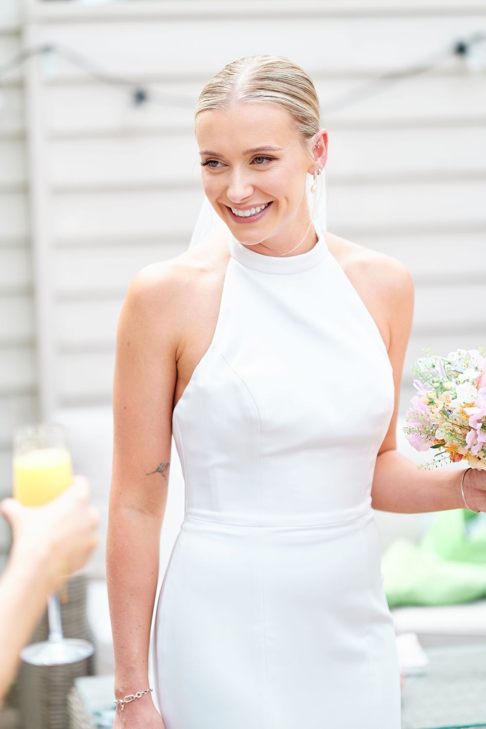 Smiling blonde bride in white halter dress holding pastel bouquet at outdoor wedding reception.
