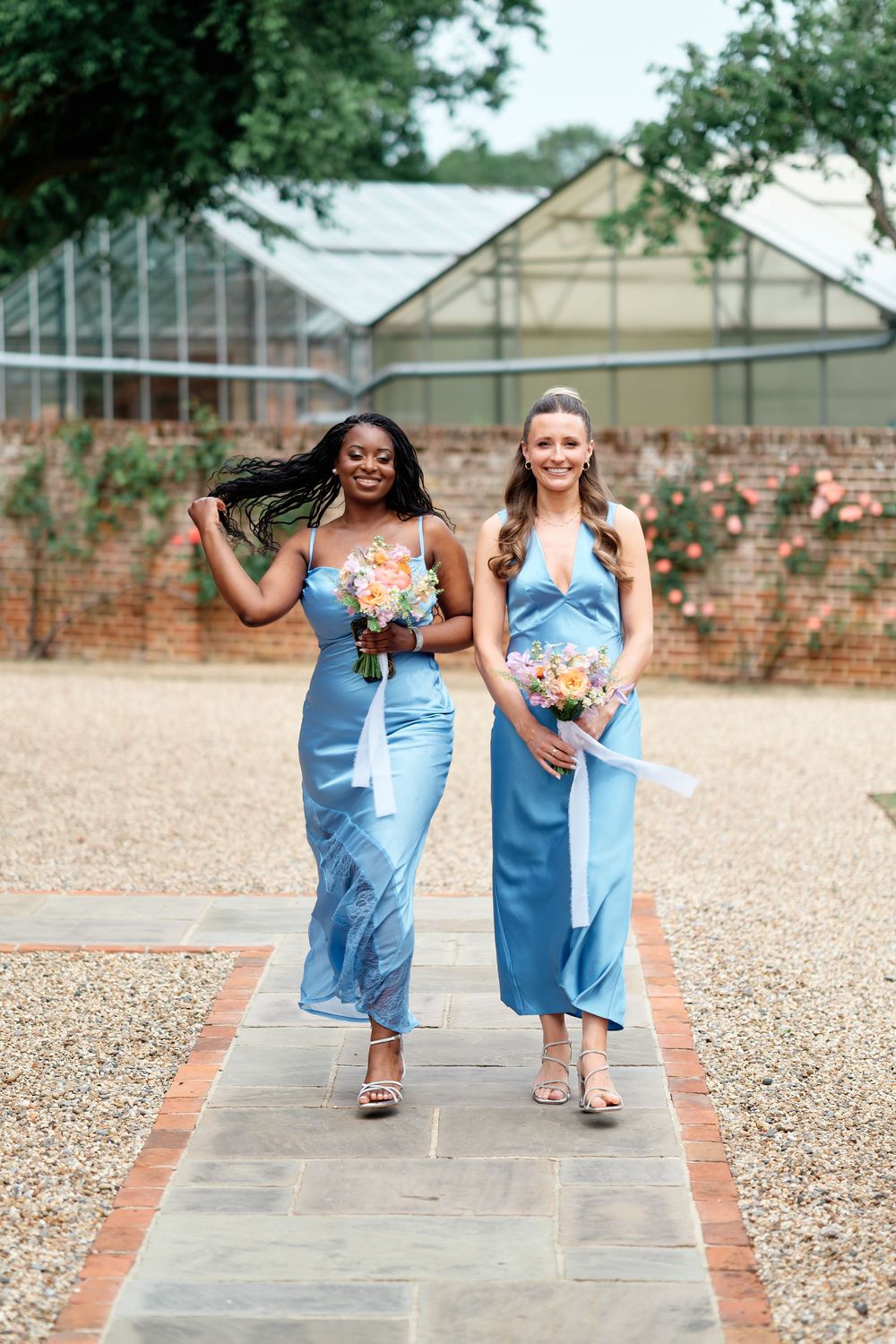Two bridesmaids in blue satin dresses walking together holding colorful bouquets at an outdoor venue.