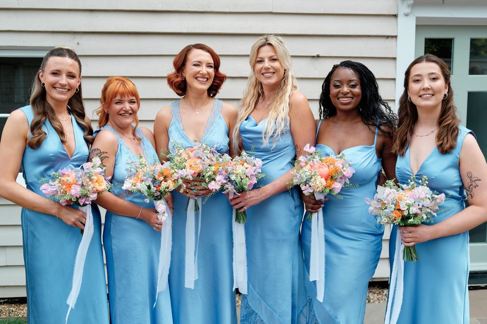 Six bridesmaids in matching light blue satin dresses holding colorful floral bouquets with white ribbons, smiling outdoors.