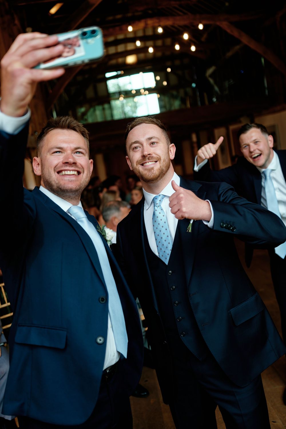 Two smiling groomsmen in navy suits take a selfie at a wedding reception venue with string lights.