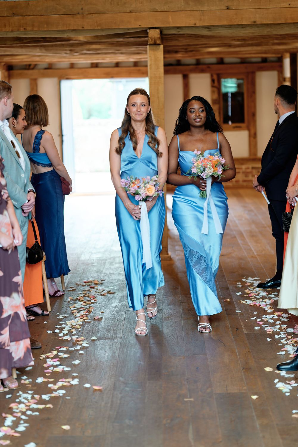 Two bridesmaids in blue satin gowns walk down the aisle holding bouquets at an elegant barn wedding.