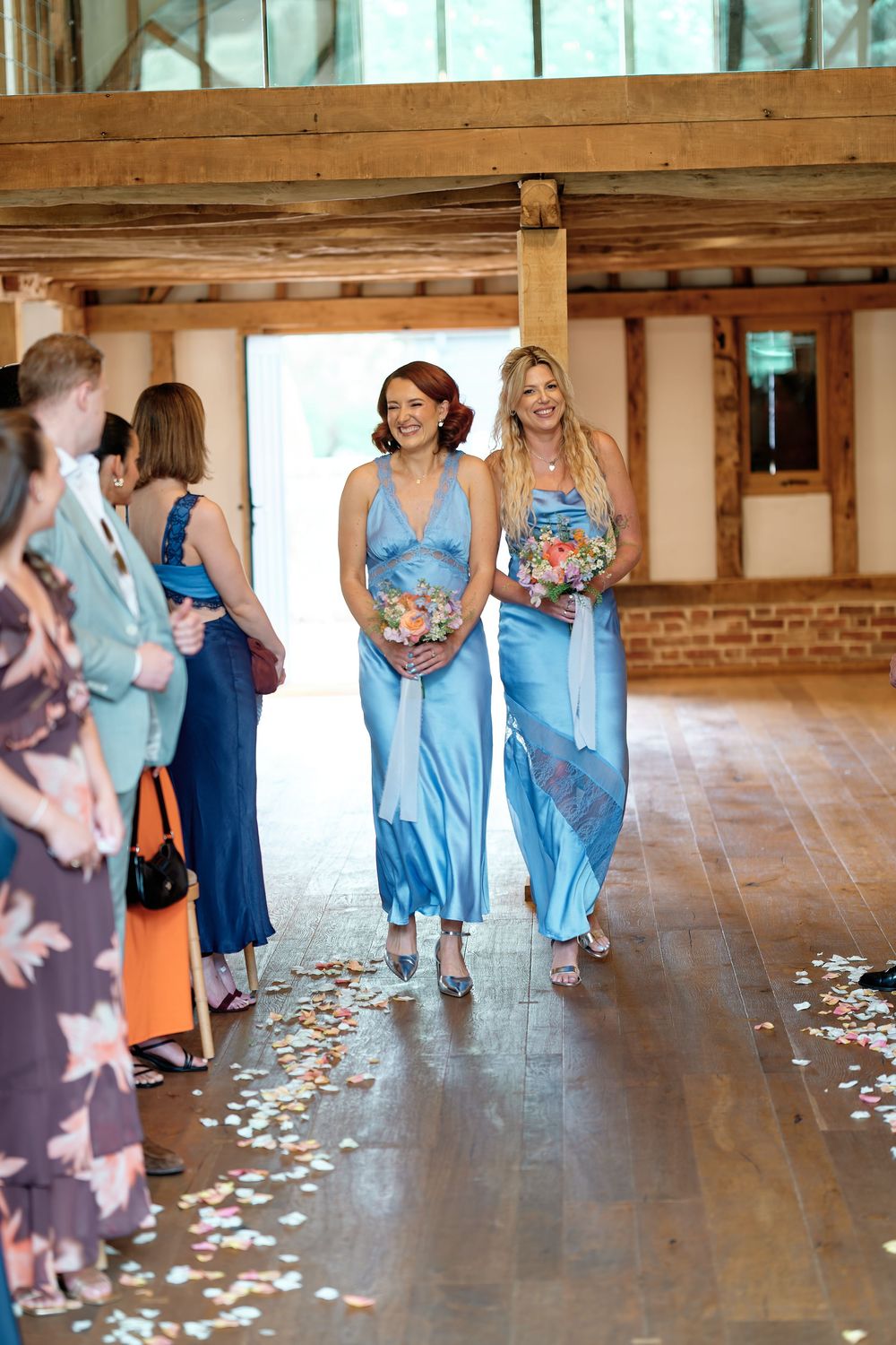 Two bridesmaids in blue satin dresses walk down a flower-petal aisle in a rustic barn wedding venue.