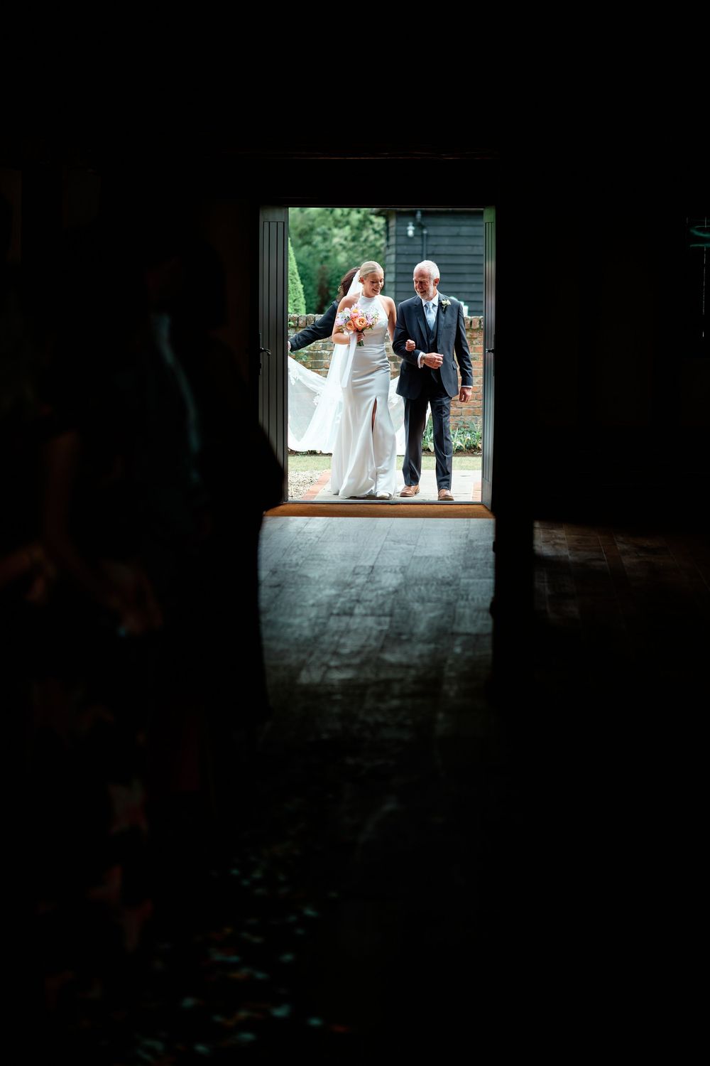 Bride and father of the bride stand in a doorway of a dark barn, ready to walk down the aisle.