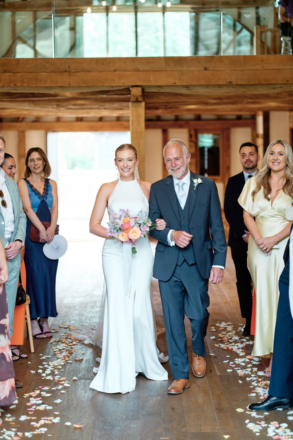 Bride in white halter gown walks down aisle with father in barn venue, holding colorful bouquet, guests lining the path.