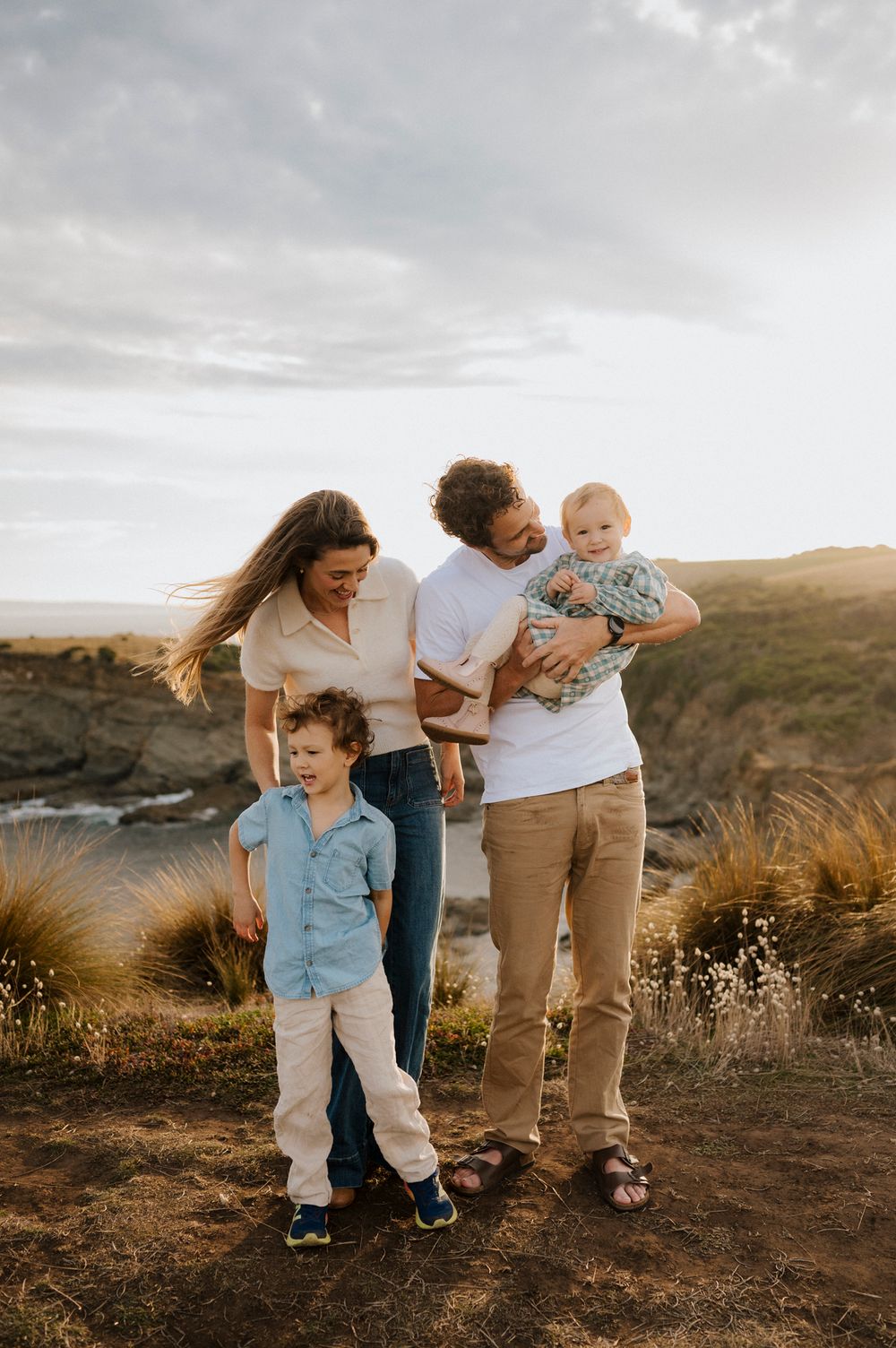 Family of four posing on coastal cliffs at golden hour, parents with two young children enjoying scenic ocean views.