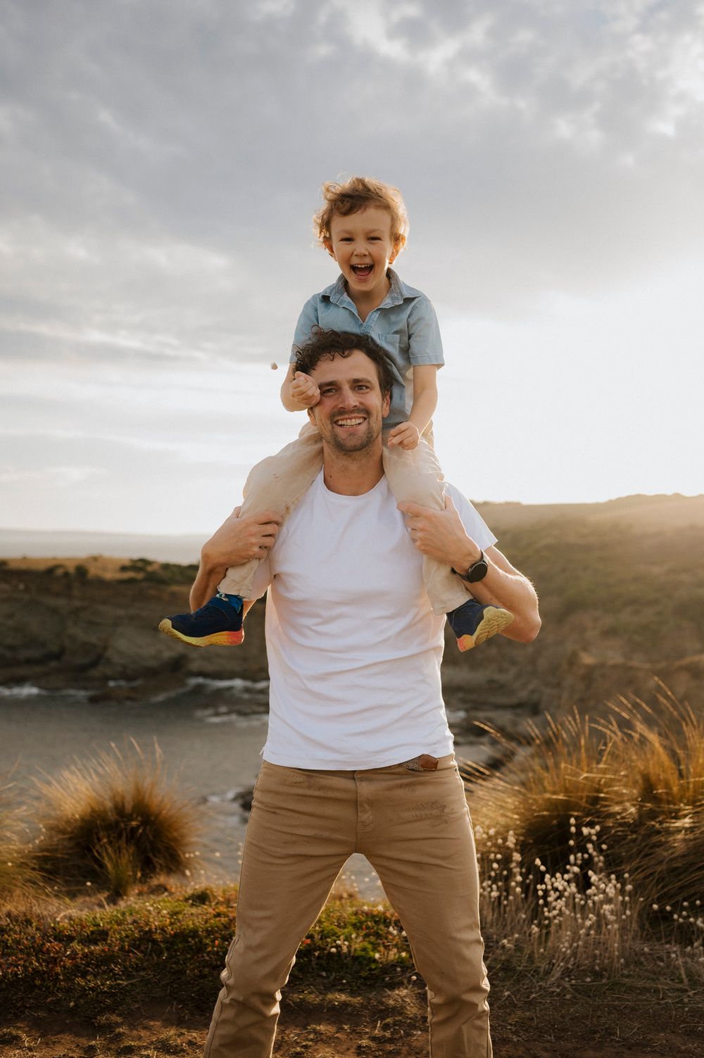 A smiling man in a white t-shirt carries a laughing toddler on his shoulders outdoors at golden hour.