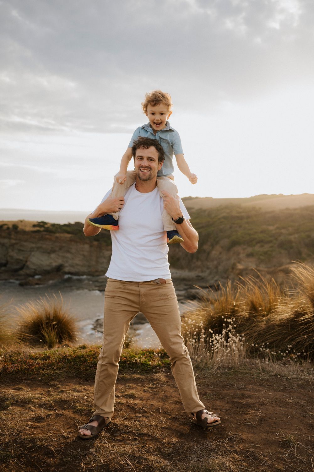 A smiling man carries a laughing child on his shoulders outdoors near coastal cliffs at golden hour.