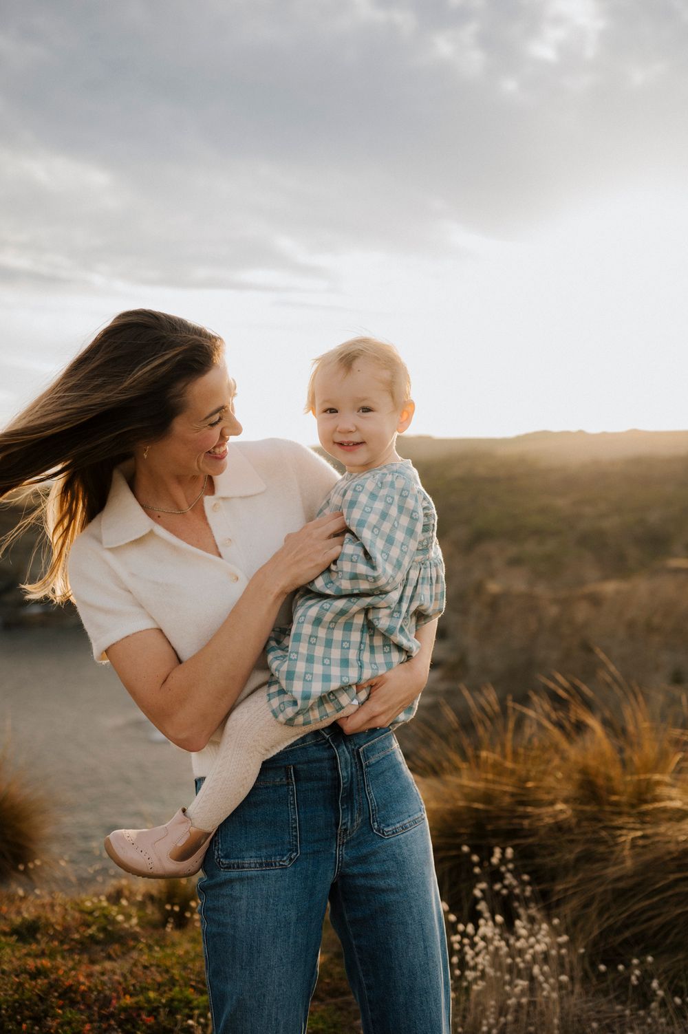 A smiling mother holds her happy toddler outdoors at golden hour, with windswept hair and a scenic landscape behind them.