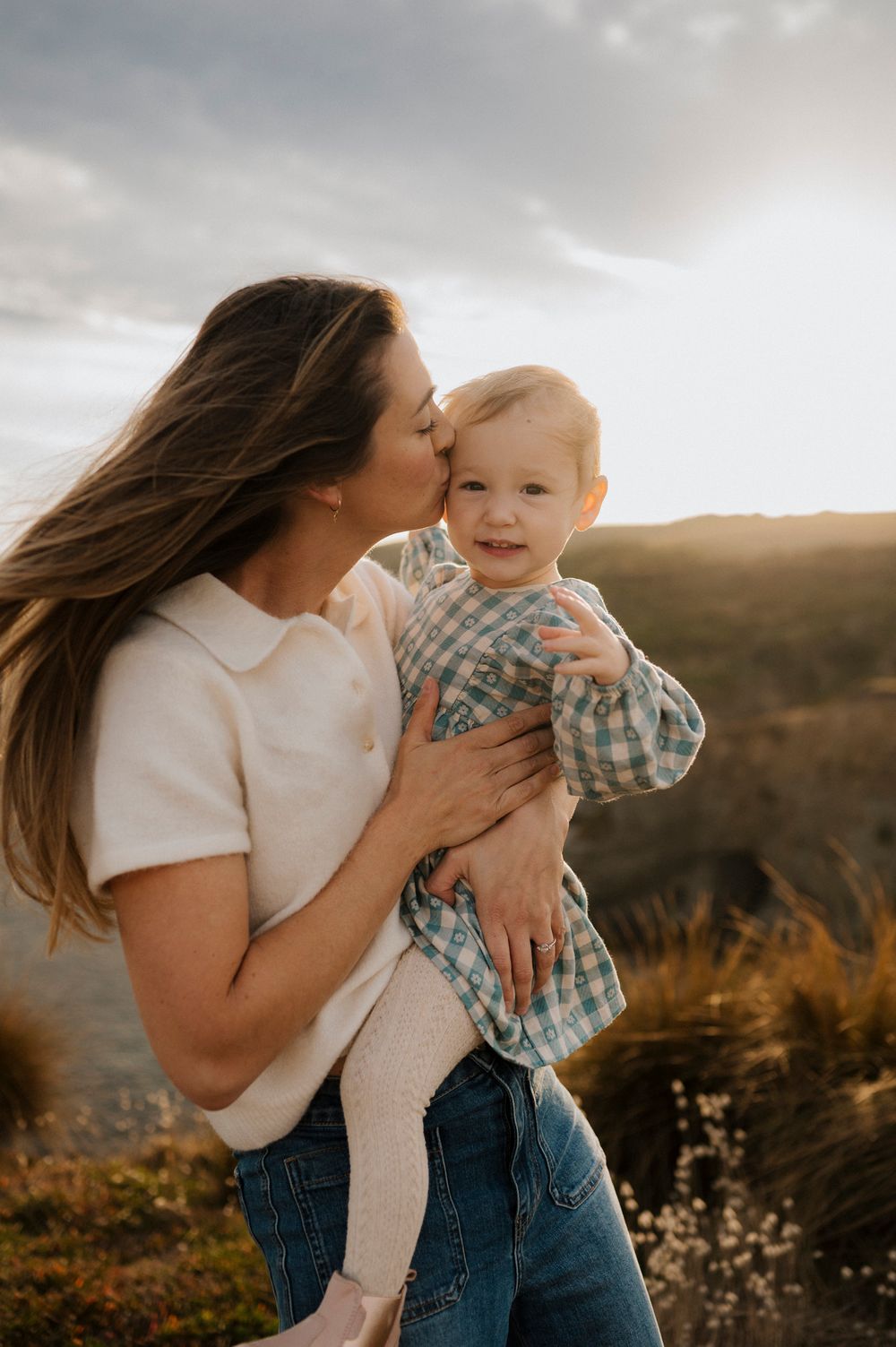 A mother kisses her smiling baby outdoors at golden hour with scenic hills in the background.