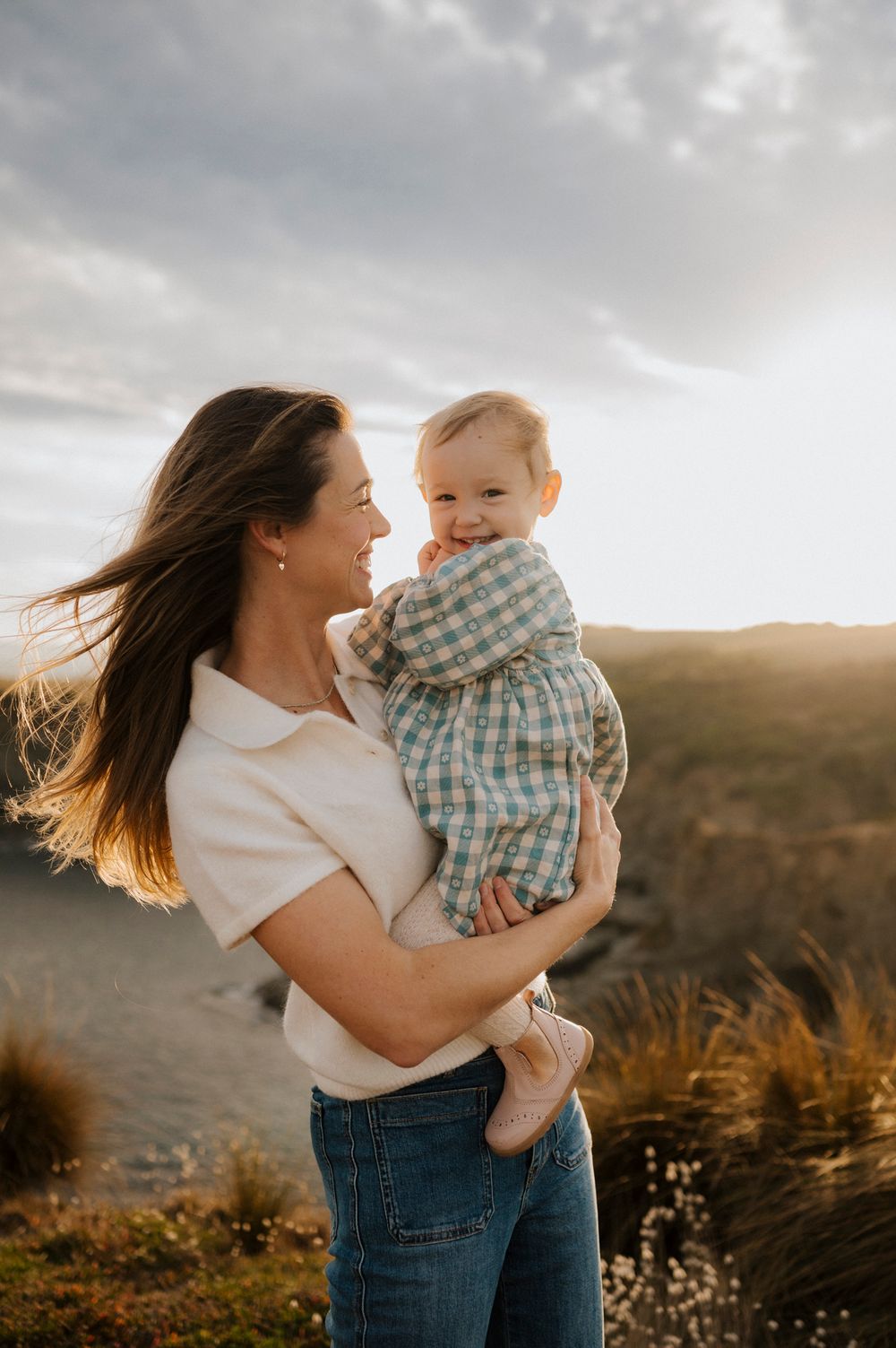 A mother holds her smiling baby outdoors at golden hour, with wind-blown hair and rolling hills in the background.