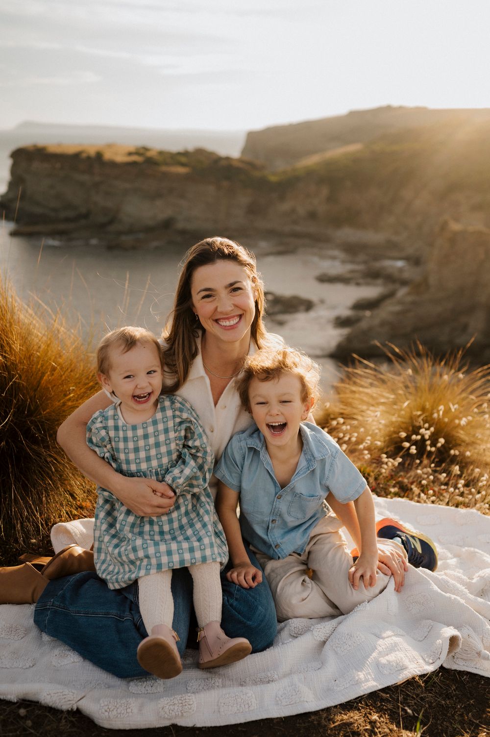 A smiling mother sits on a white blanket outdoors with her two laughing young children, coastal cliffs at golden hour.