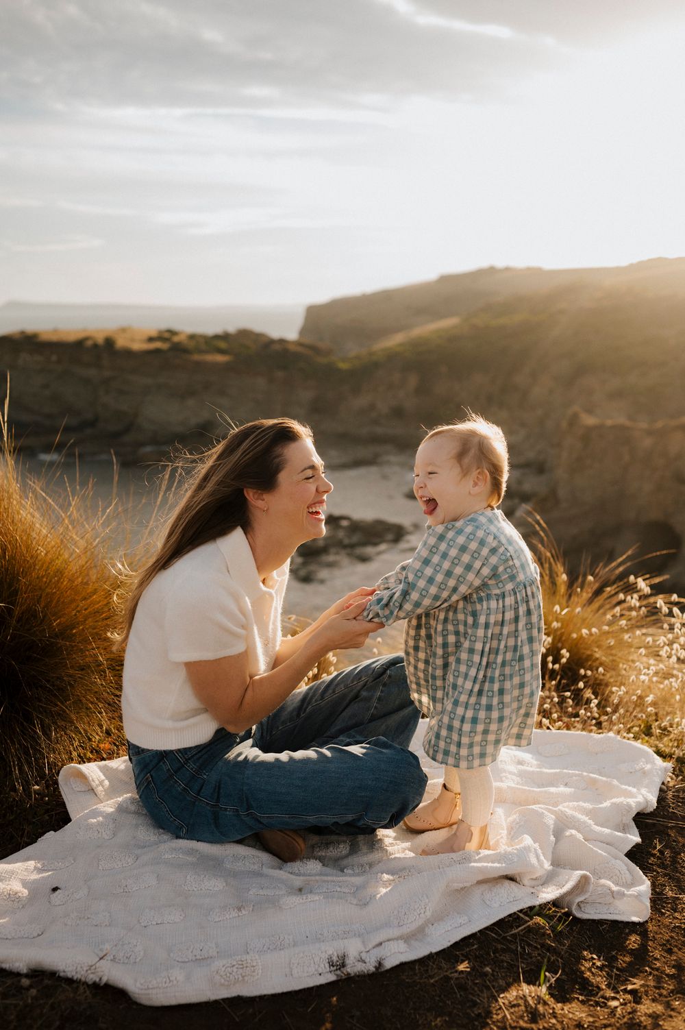 A mother and baby share a joyful moment on a blanket at golden hour near coastal cliffs.