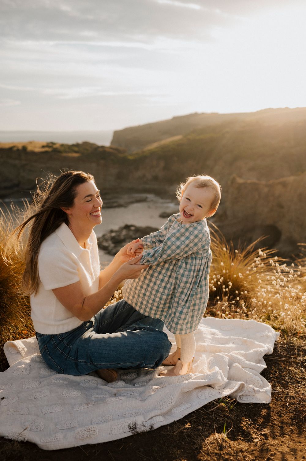 A smiling mother sits on a blanket outdoors at golden hour, holding her laughing baby standing on a scenic clifftop.