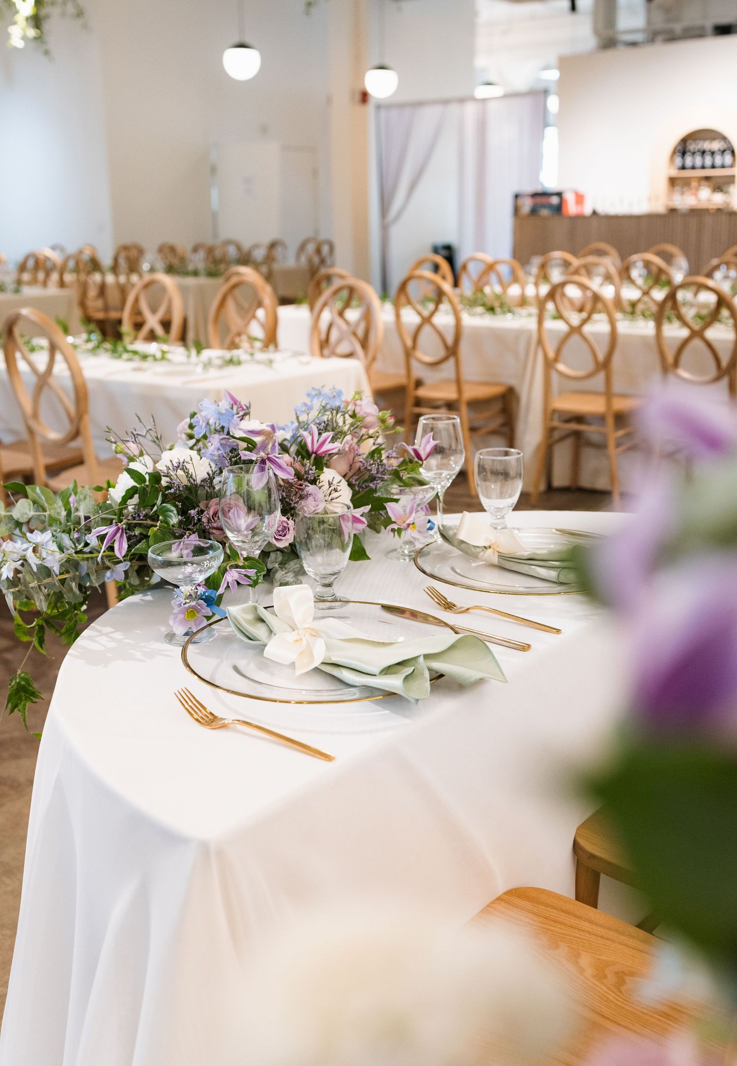 Elegant wedding reception tables with white linens, gold cutlery, and purple floral centerpieces in a bright venue.