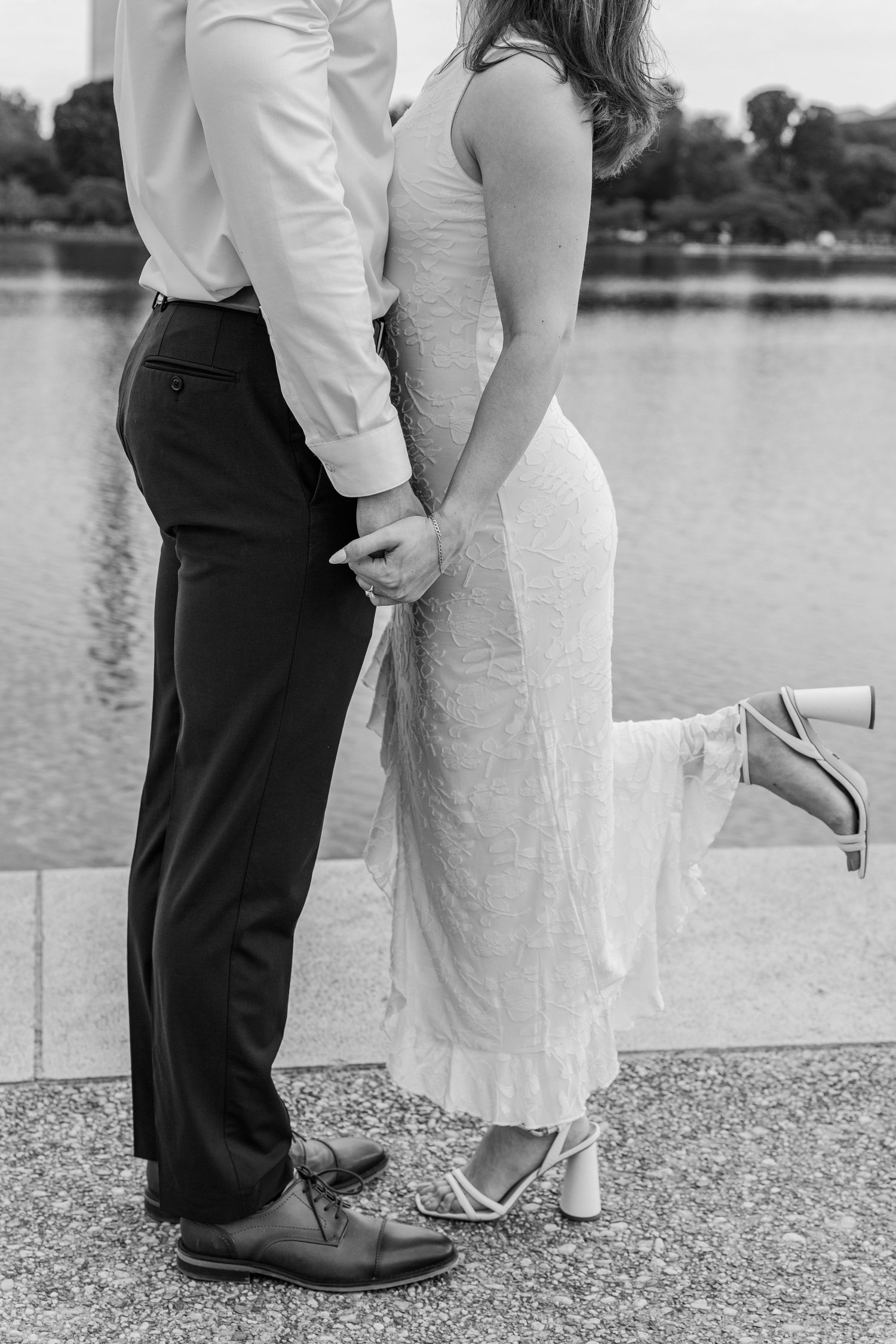 Black and white photo of a couple holding hands by a lake, woman in white dress lifting her heel, man in dress pants.