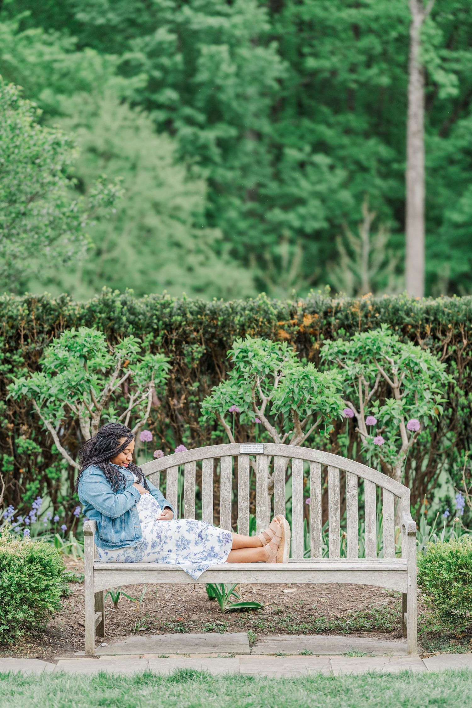 Pregnant woman in denim jacket and floral dress relaxing on wooden garden bench surrounded by lush green hedges.
