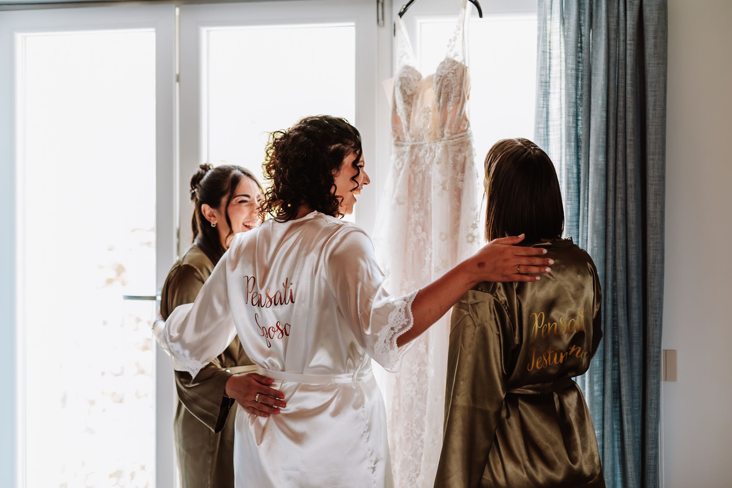 Bride and bridesmaids in robes admiring a lace wedding dress hanging by a bright window before the ceremony.