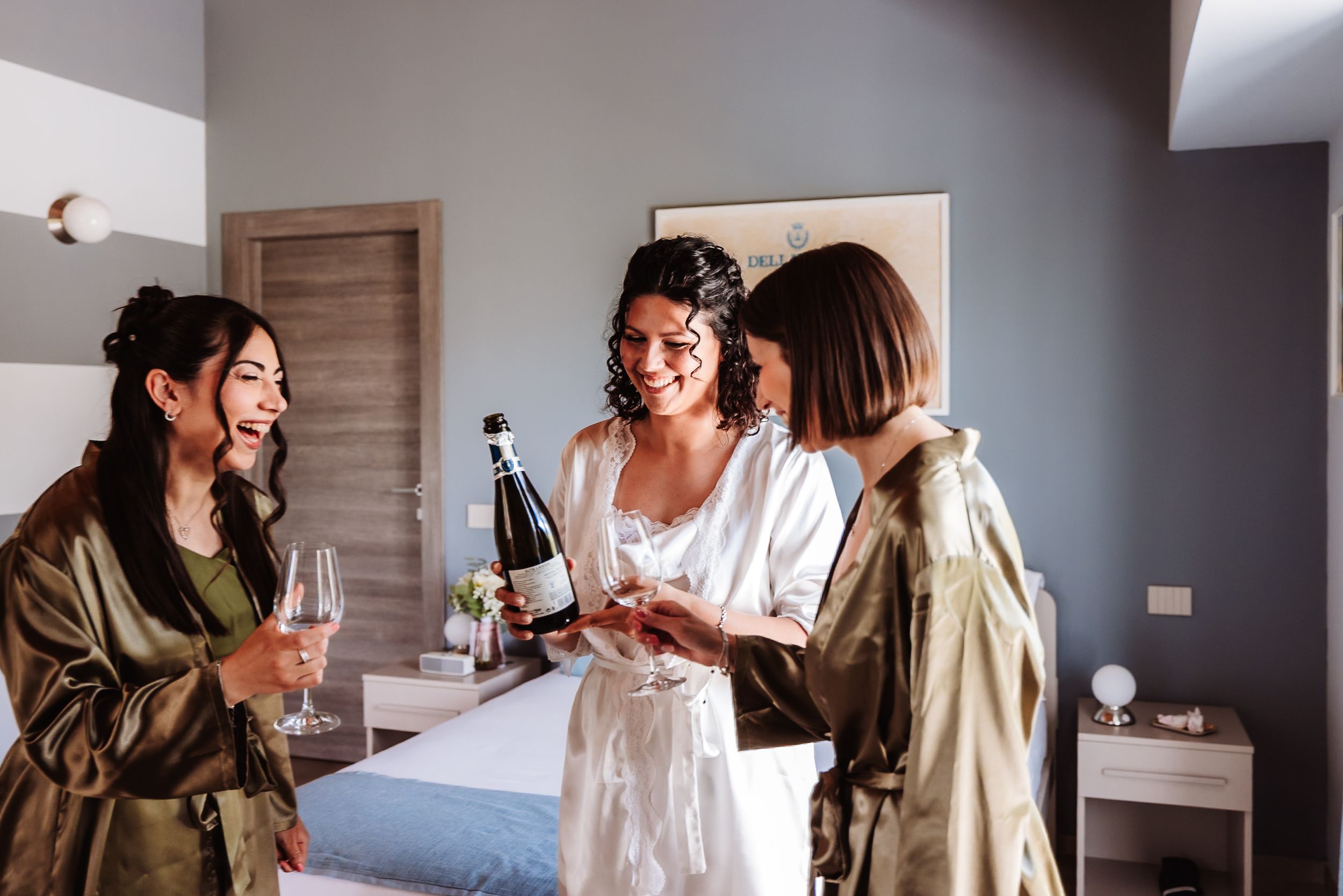 Three women in robes celebrate with champagne bottles and glasses in a bright bedroom, likely a bridal party getting ready.