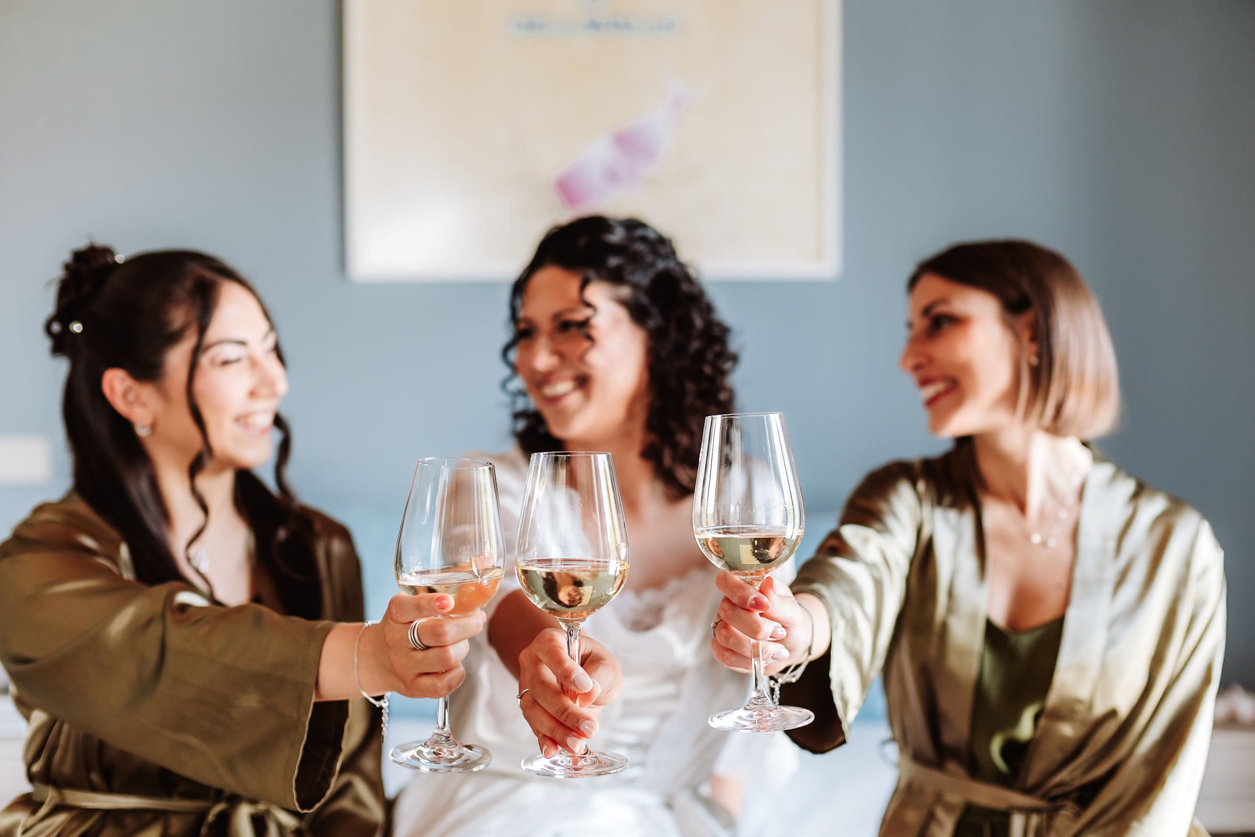Three women in satin robes toasting with white wine glasses, smiling and celebrating at a bridal party gathering.