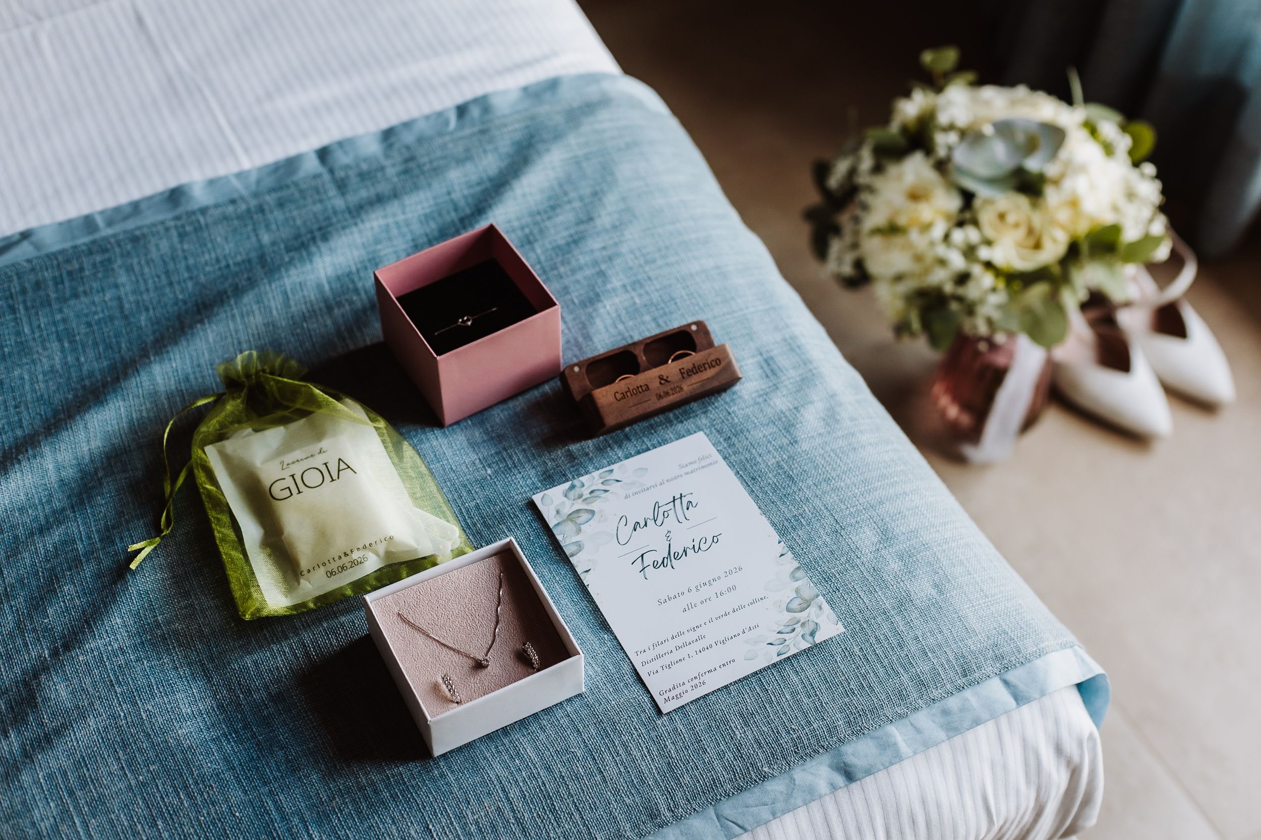 Wedding flat lay with perfume, jewelry boxes, necklace, invitation, bridal bouquet, and white heels on blue bed.