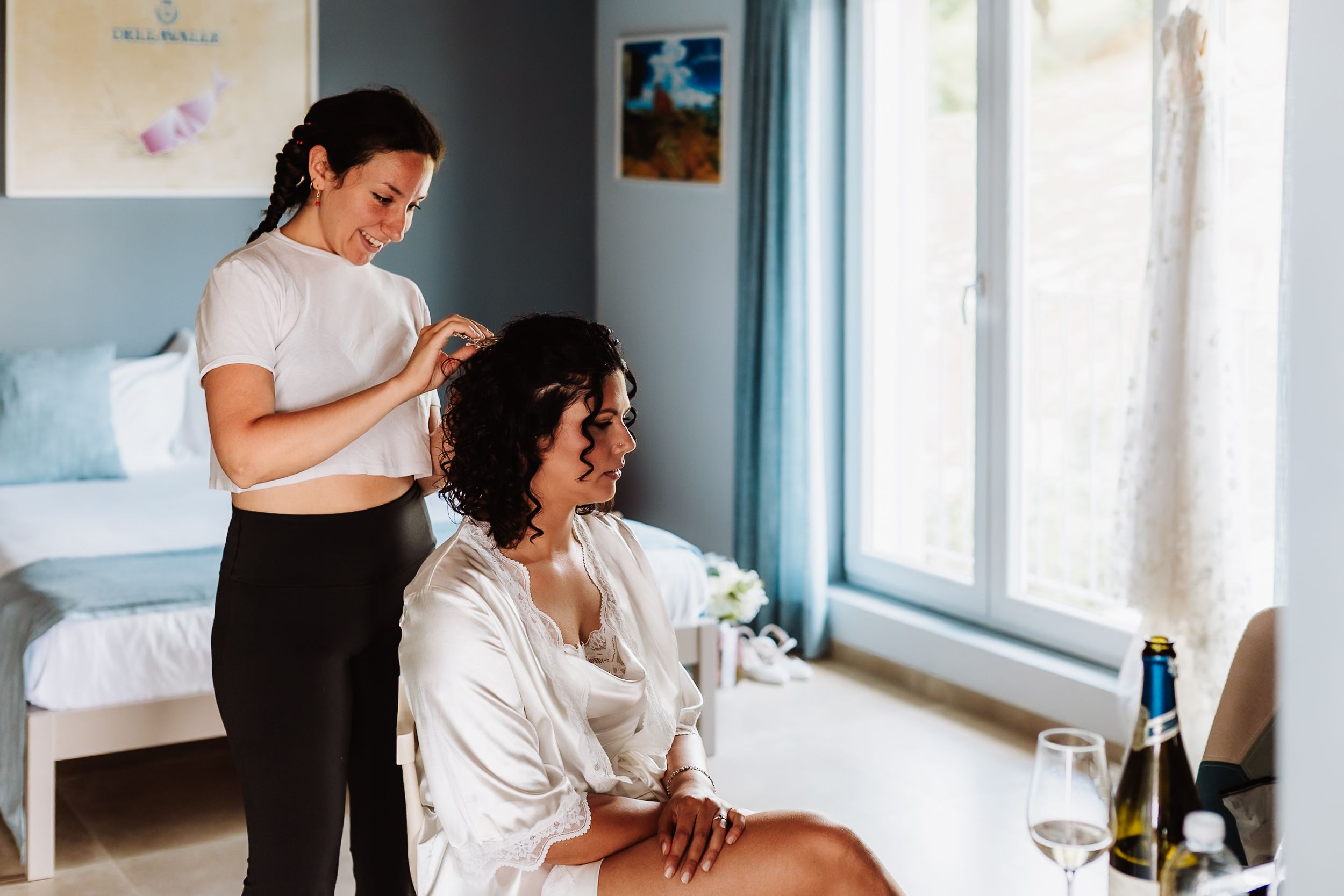 A hairstylist styling a bride's hair in a bright room with champagne nearby on a wedding morning.