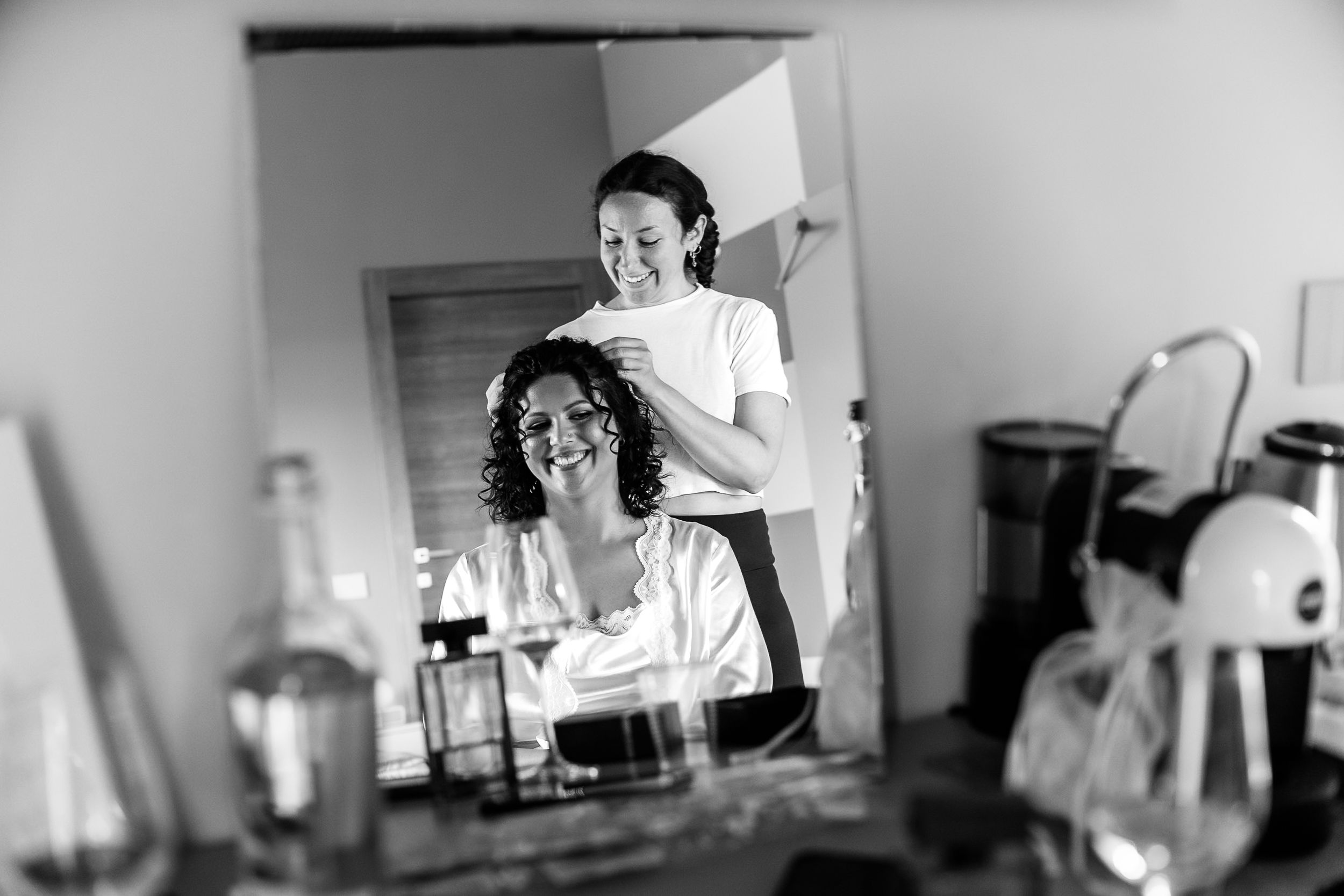 Black and white photo of a hairstylist styling a smiling bride's curly hair while reflected in a mirror.