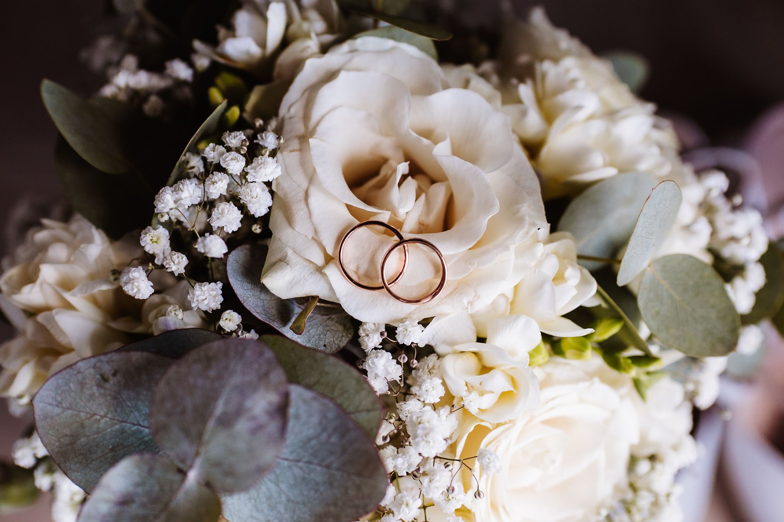 Two gold wedding rings rest on a bridal bouquet of white roses, baby's breath, and eucalyptus leaves.