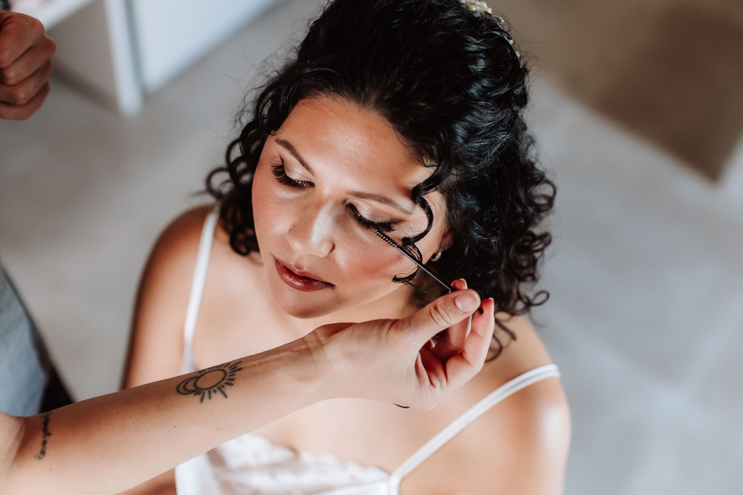 A bride with curly dark hair gets her makeup applied, showcasing a floral tattoo on her arm.