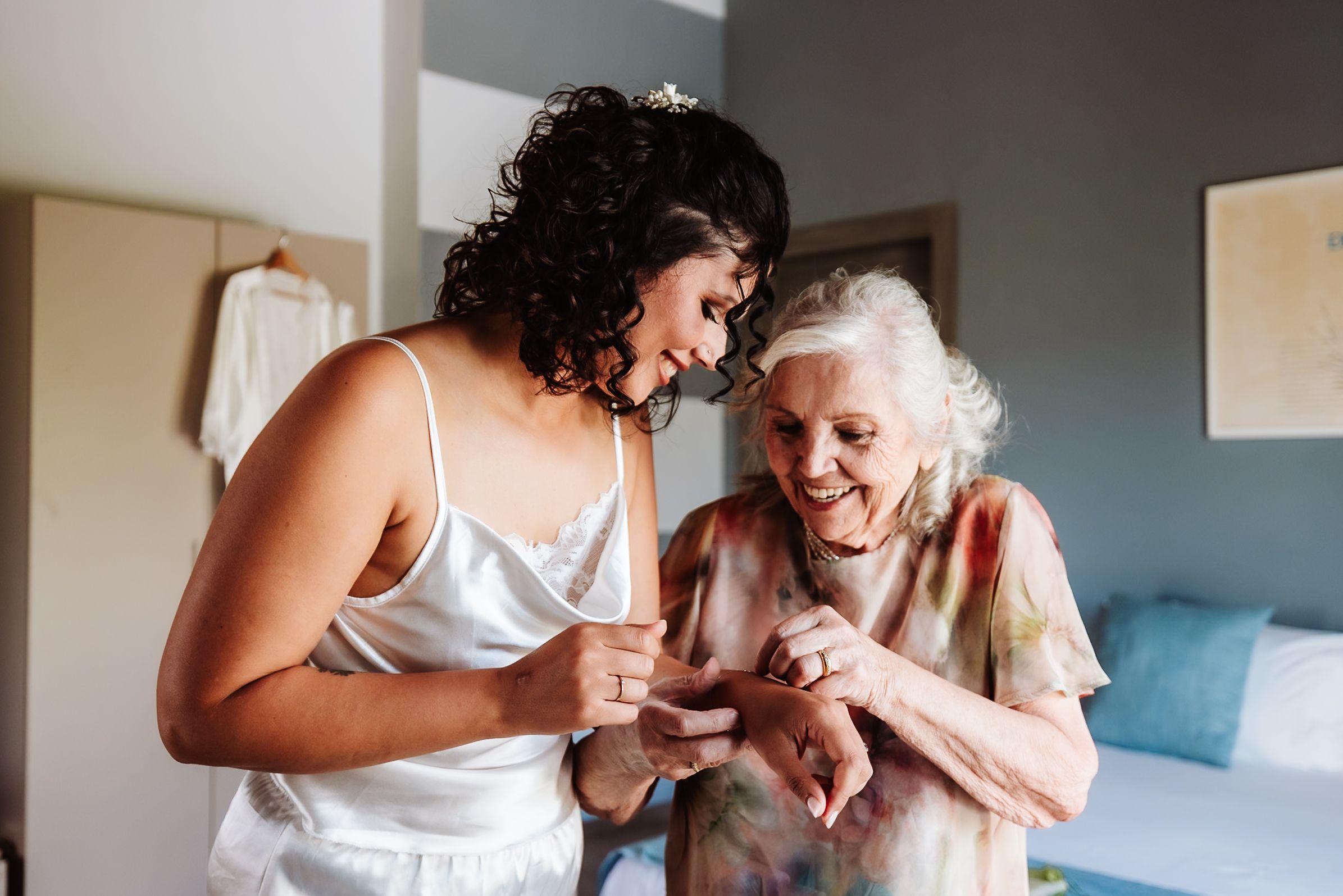 Bride in white slip dress shares a joyful moment with elderly woman examining jewelry during wedding preparations.