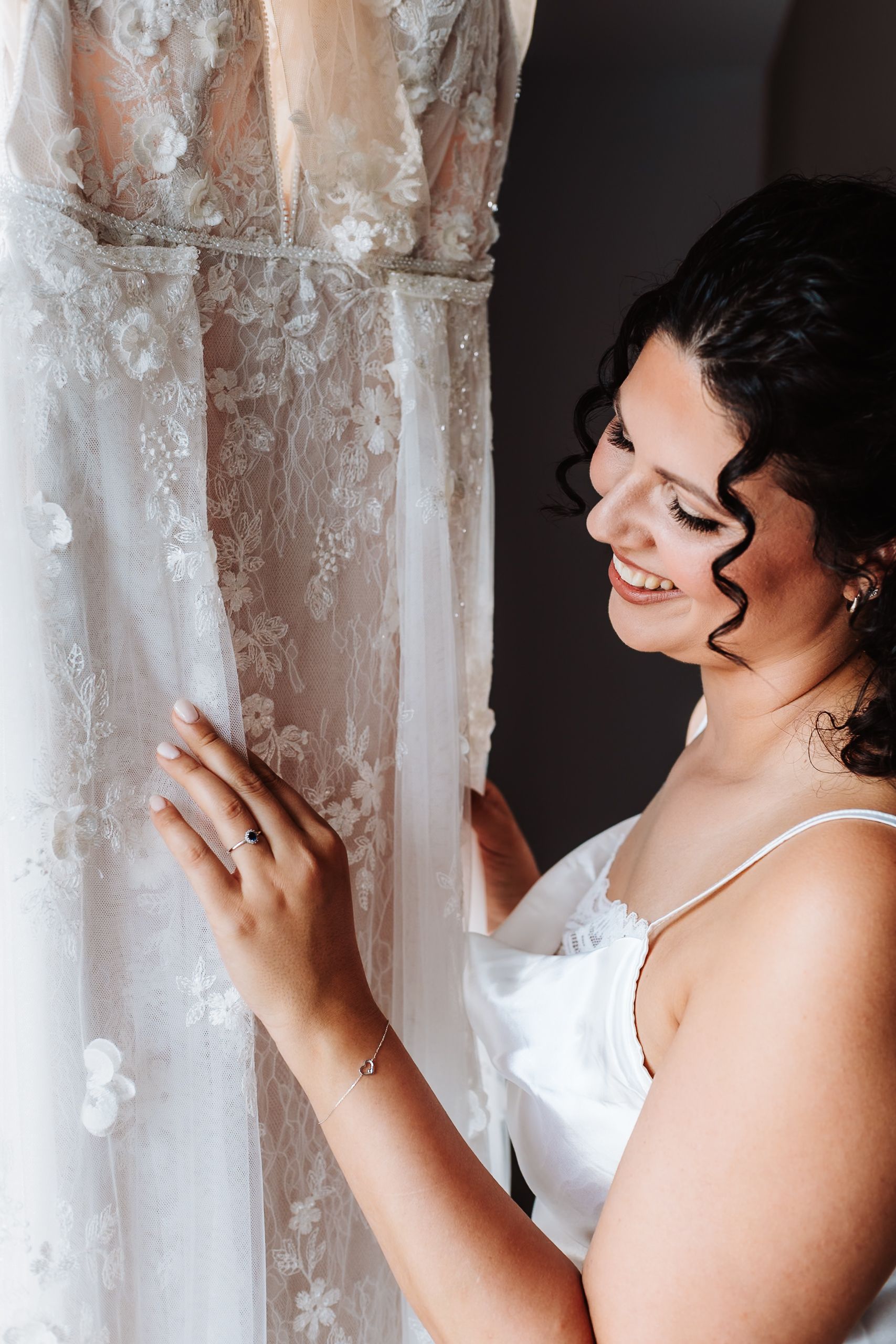 Smiling bride in white slip dress admires her lace floral wedding gown hanging up before the ceremony.
