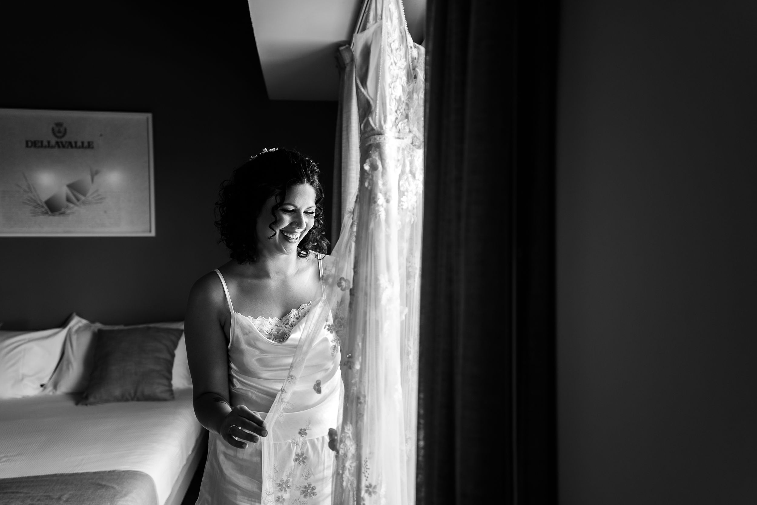 Black and white photo of a smiling bride in a wedding dress standing by a window in a hotel room.