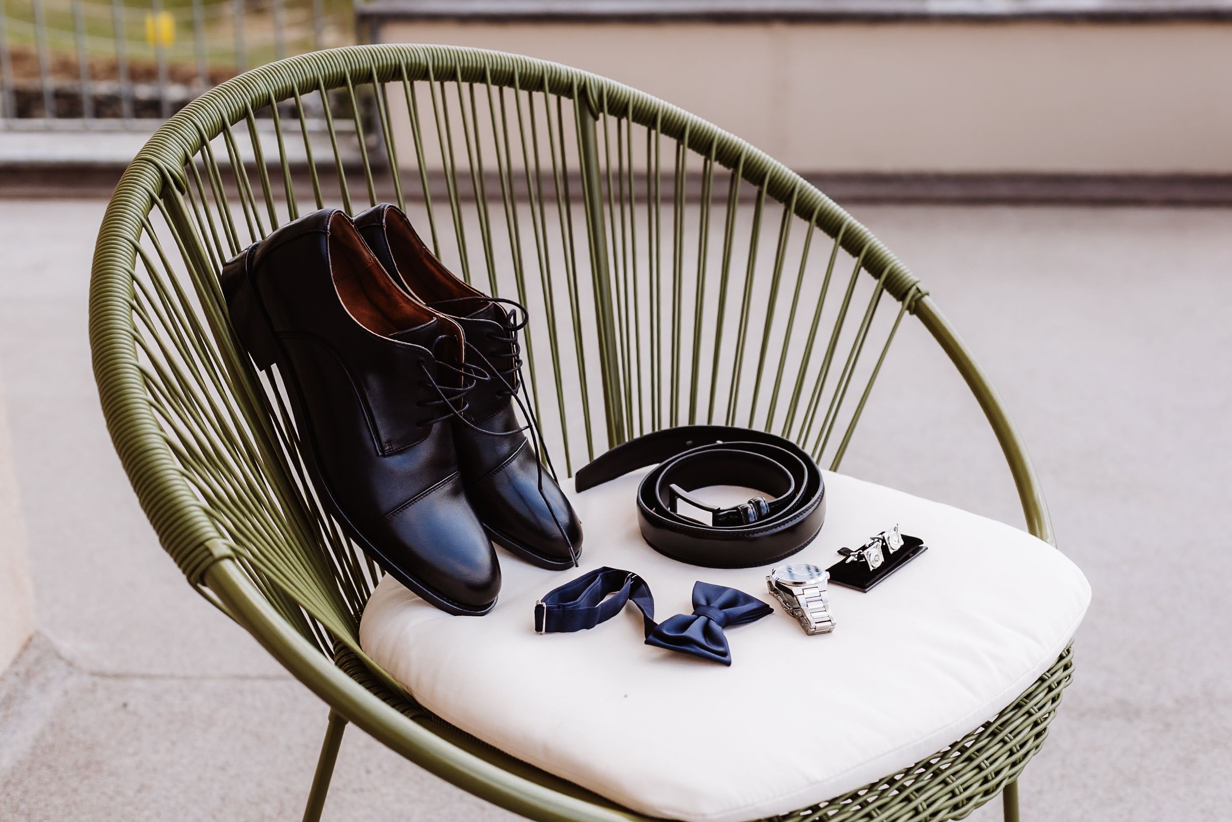 Black dress shoes, belt, bow tie, and cufflinks arranged on a green wicker chair for a groom's wedding accessories flat lay.